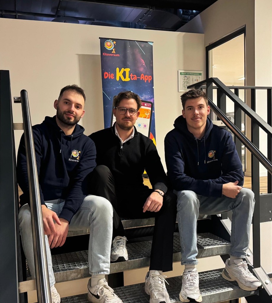 Das Gründerteam von Kitaversum – drei Gründer sitzen auf einer Treppe vor dem Kitaversum-Banner